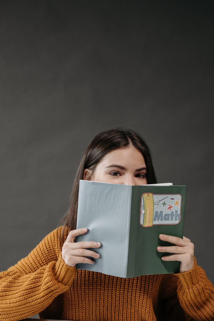 hero-img-02 A teenage girl holds a math book while partially hidden, highlighted against a gray background.